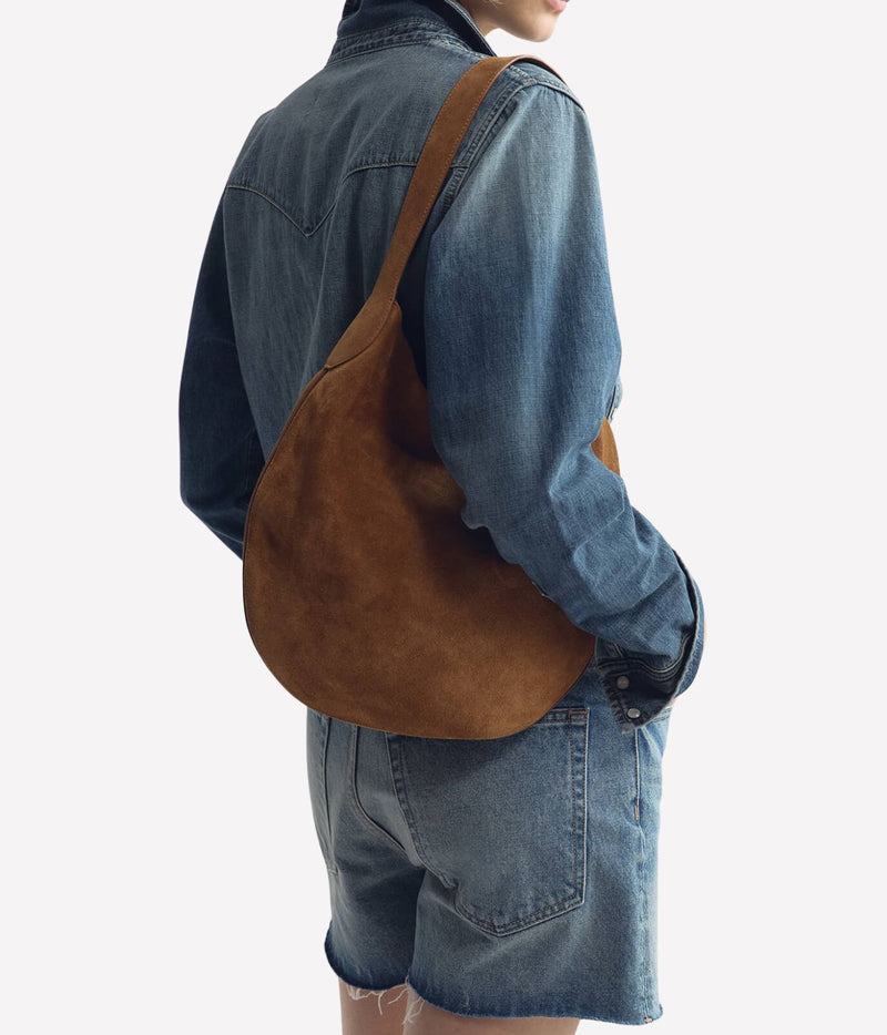 A person wearing a blue denim shirt and shorts, shown from the back and side, carrying a large brown suede shoulder bag over the shoulder against a plain light background.