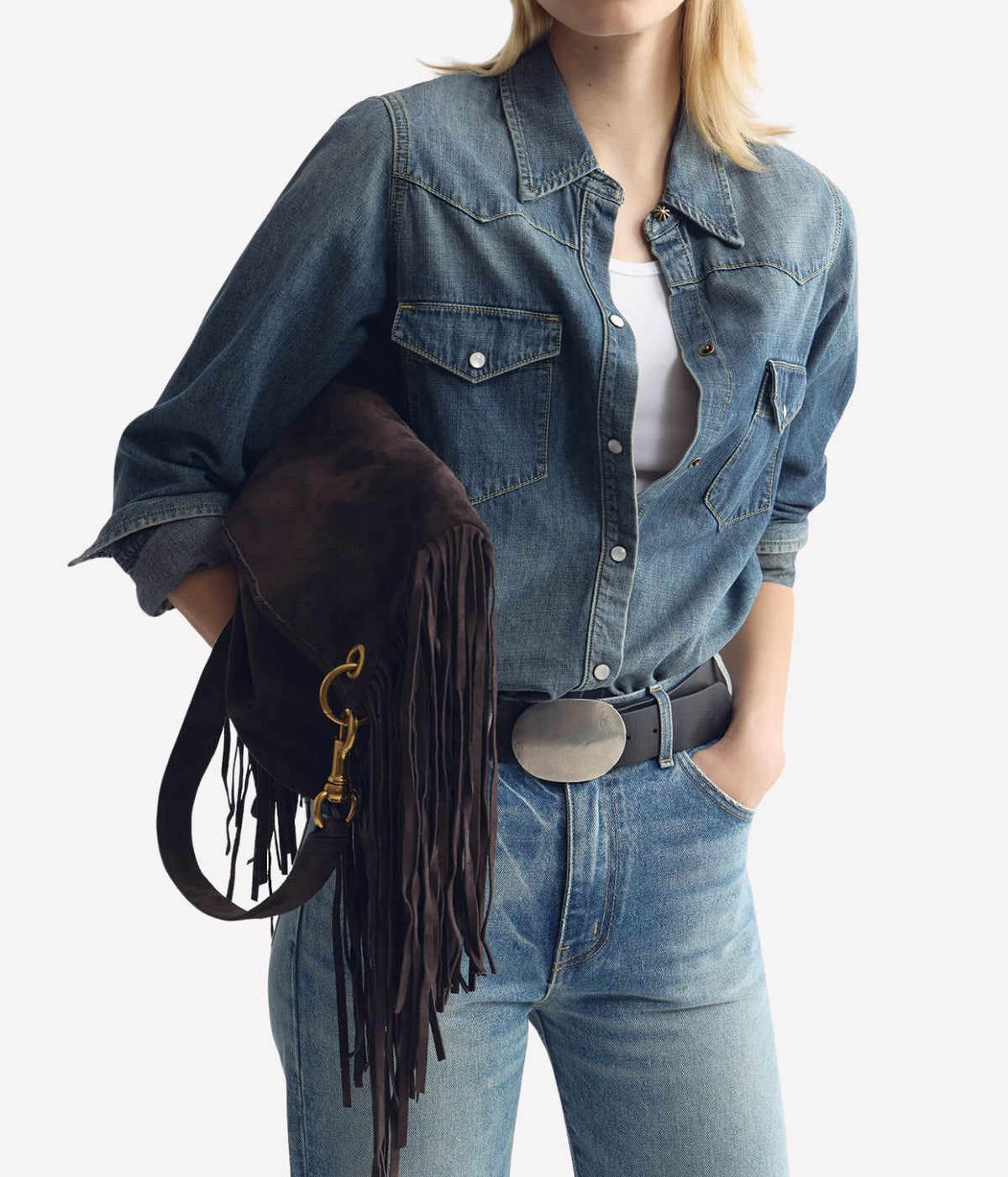 A woman wearing a blue denim shirt and jeans, holding a dark brown suede fringe shoulder bag with gold hardware against a light gray background.