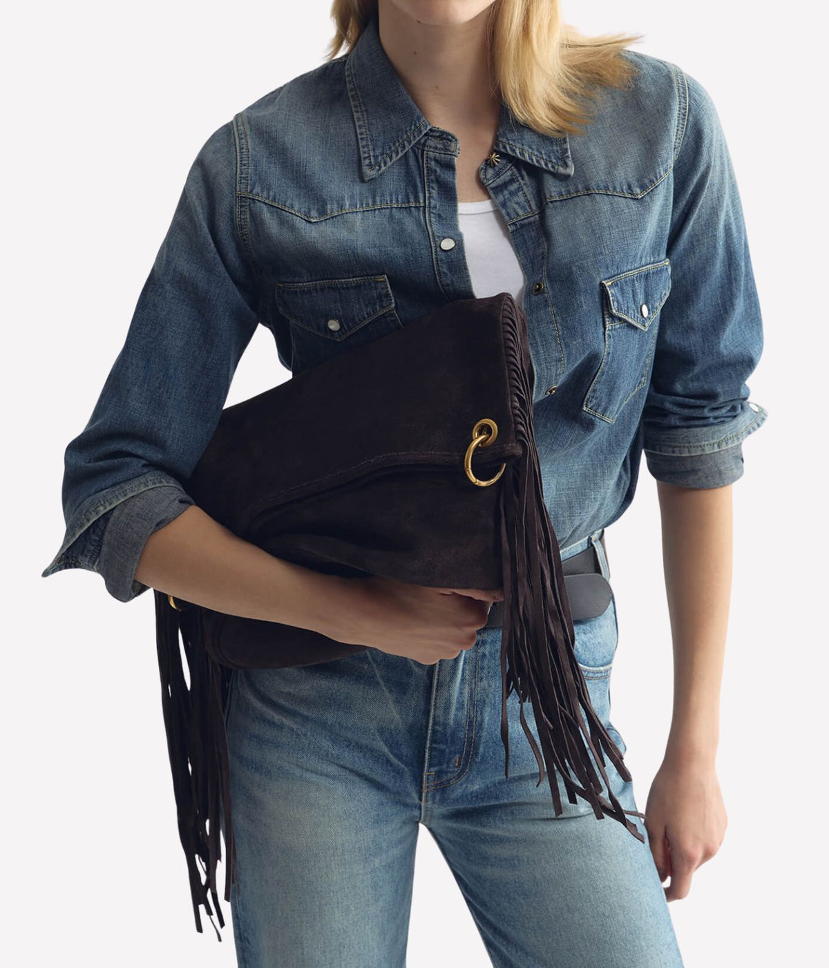 A model wearing a blue denim shirt and jeans holds a dark brown suede fringe clutch bag with a gold-tone ring clasp against a light gray background.