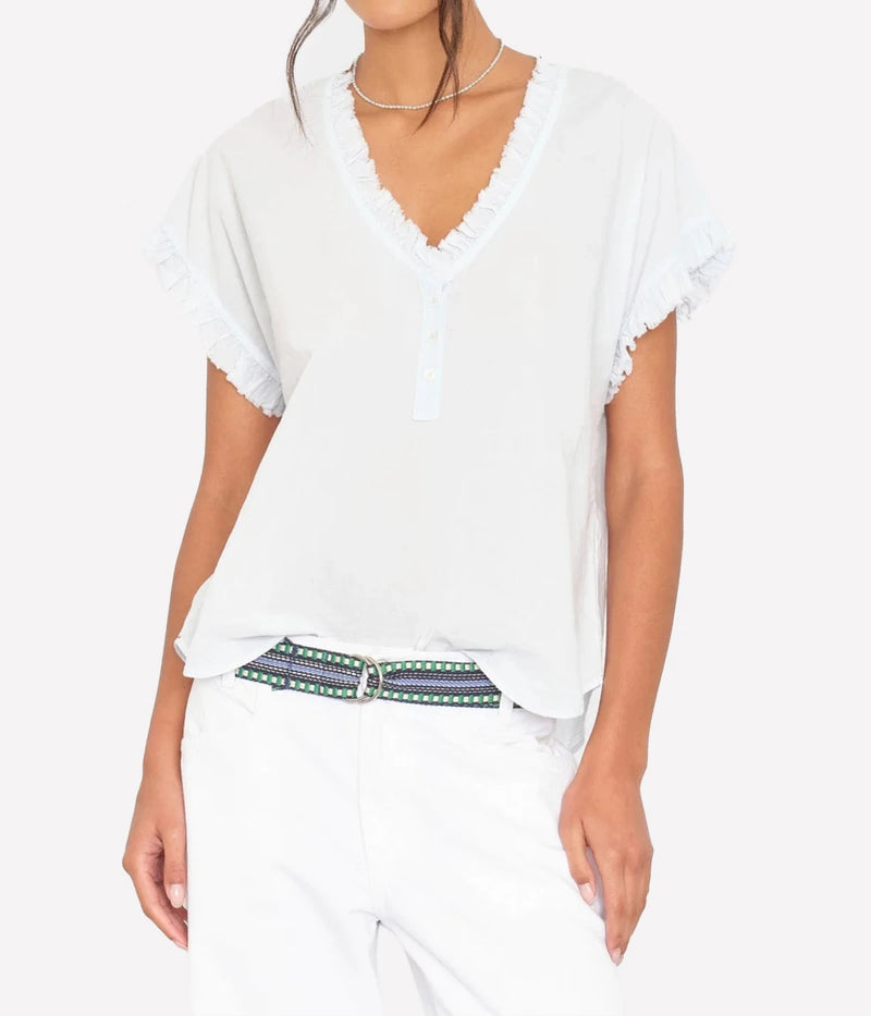 Model wearing the Xirena Darcy Top in Skylight loosely tucked into white jeans with a striped woven belt and a fine silver chain necklace, showing the ruffled V-neckline