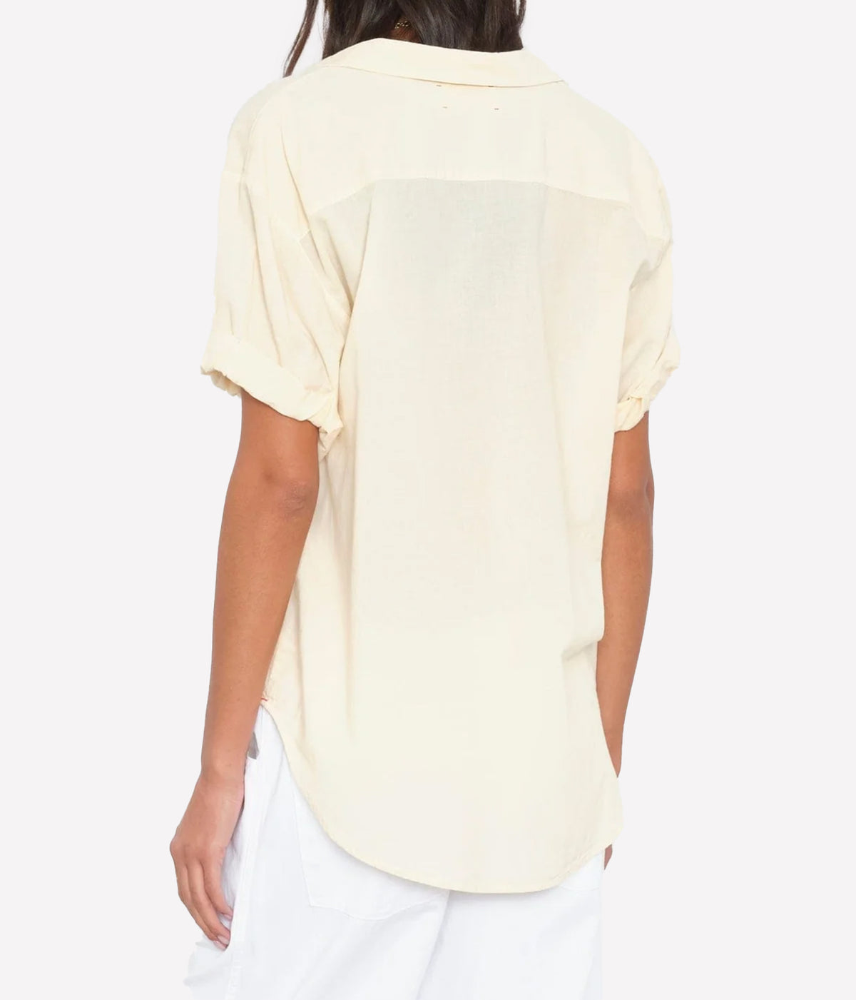 Back view of a model wearing the Xirena Channing Shirt in Oysterette with white pants, showing the back yoke, rolled sleeves and curved shirttail hem