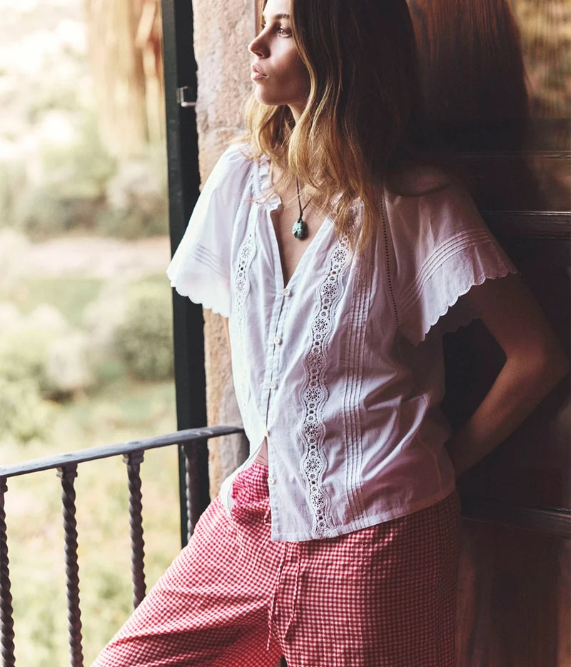 Woman wearing a white button-front blouse with flutter sleeves and red gingham trousers, standing beside a window.