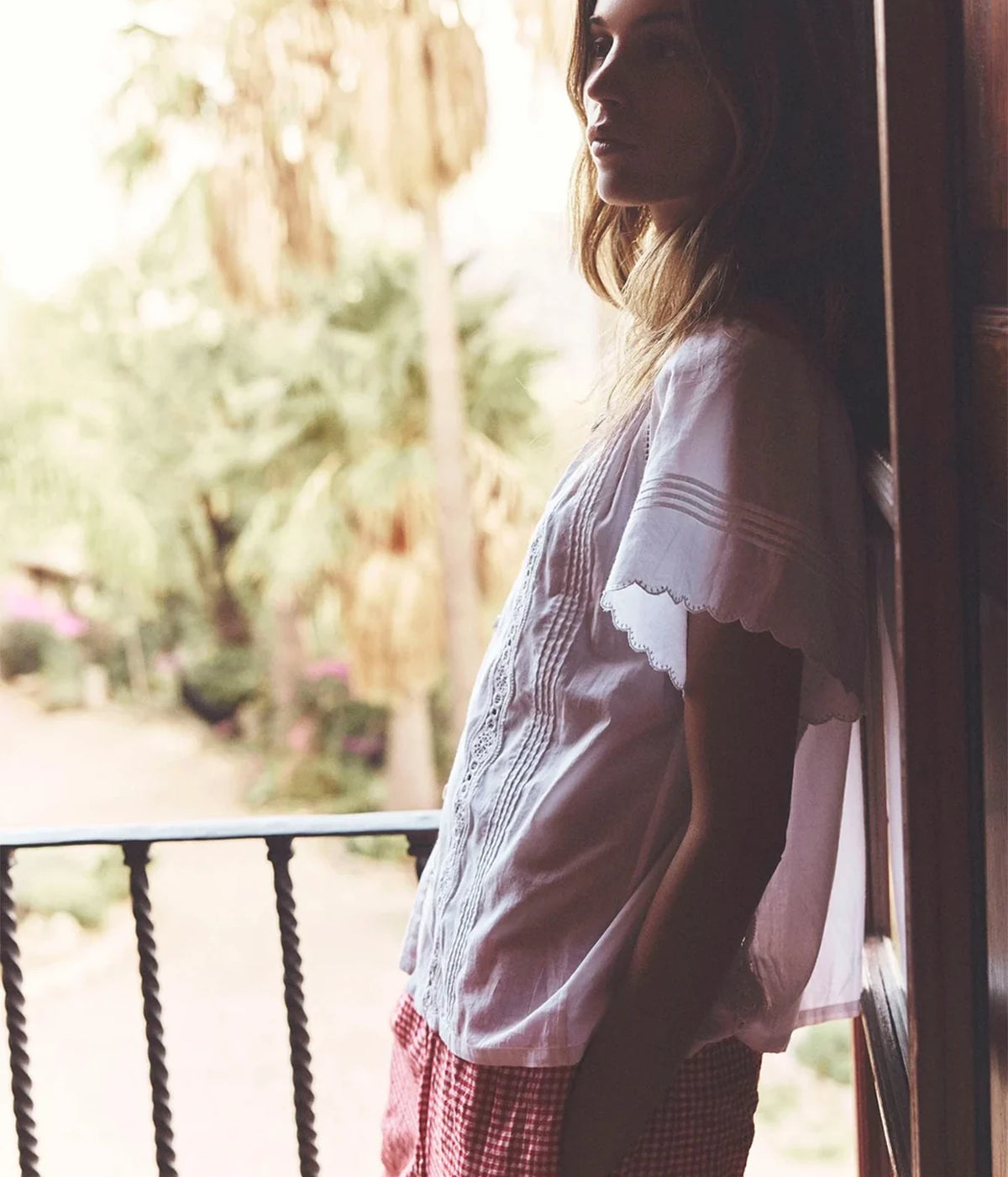 A woman wearing a white embroidered short-sleeve blouse and red-and-white gingham trousers, leaning against a dark wooden door on a balcony