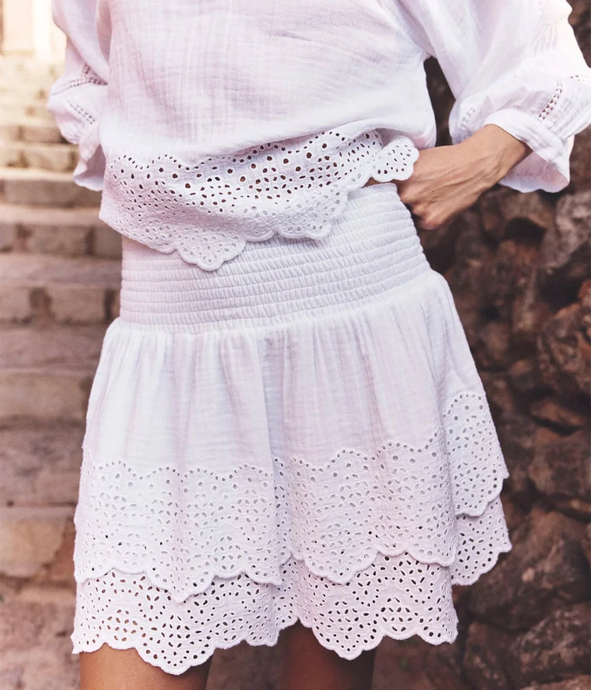 A close-up of a person wearing the white Roxana blouse and matching Xirena Rima Skirt in White with scalloped eyelet trim, standing outdoors against a rustic stone wall.
