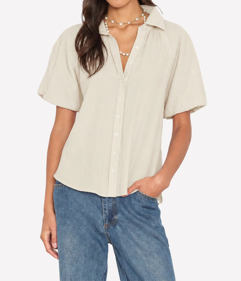 Model wearing the Xirena Lyla Shirt in Parchment open at the neck over mid-wash blue jeans, with a pearl beaded necklace and one hand in her pocket